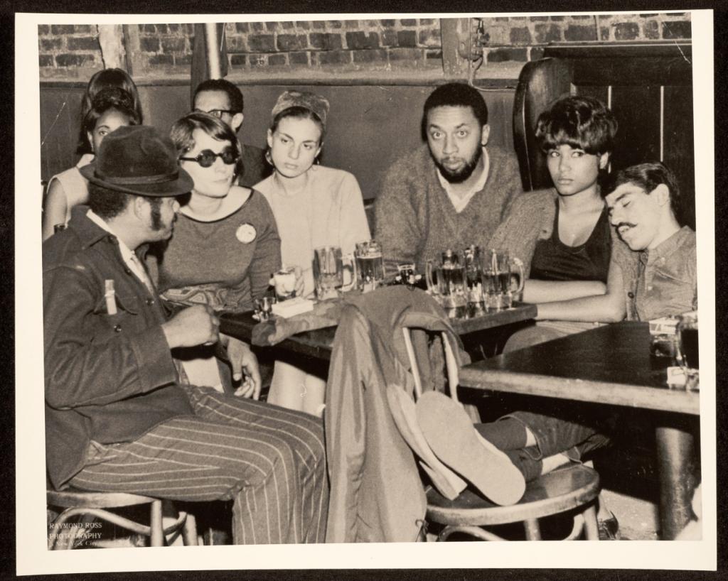 A group of siz people sit around a table at a table. On the left, three are engaged in a conversation, the man in the center is looking at the camera, and the man on the right is sleeping on the shoulder of the woman next to him, who is looking off to the side.