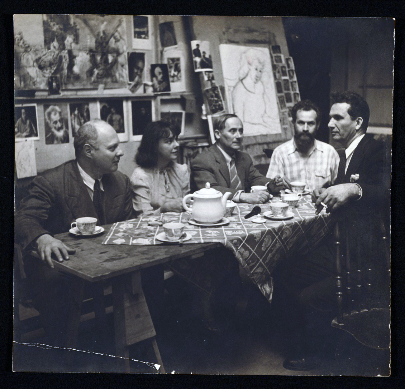 Four men and one woman sit at a table, drinking tea and talking in an artist's studio.