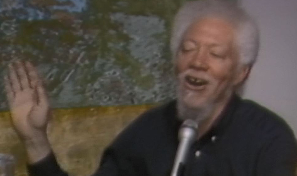 Benny Andrews sits at a table with a microphone, cups, and a pitcher of water with a yellow, green, and blue abstract painting hanging behind him. He is wearing a black button down shirt and raising his hands while talking. 