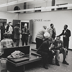 Black and white photograph, taken during the summer of 1969, shows attendees at an Cinque Gallery exhibit opening, including cofounders Norman Lewis (standing in a black suite at far right) and Romare Bearden  (farther left, talking to an unidentified man).