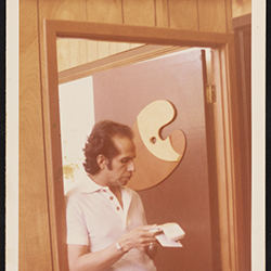 Color photograph of Mel Casas standing in a doorway looking at papers, circa 1977