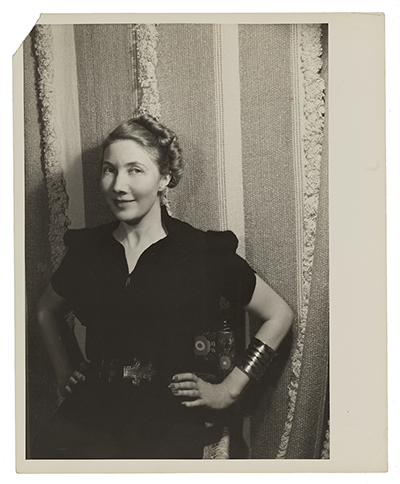 Woman in dark colored dress wearing a large cuff bracelet, photographed against one of her textile designs, which consists of various colors and textures in thick stripes.