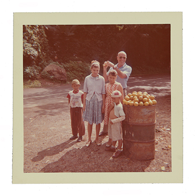 Color photograph of Emilio Sanchez posing with children in Puerto Rico next to a barrel of fruit.
