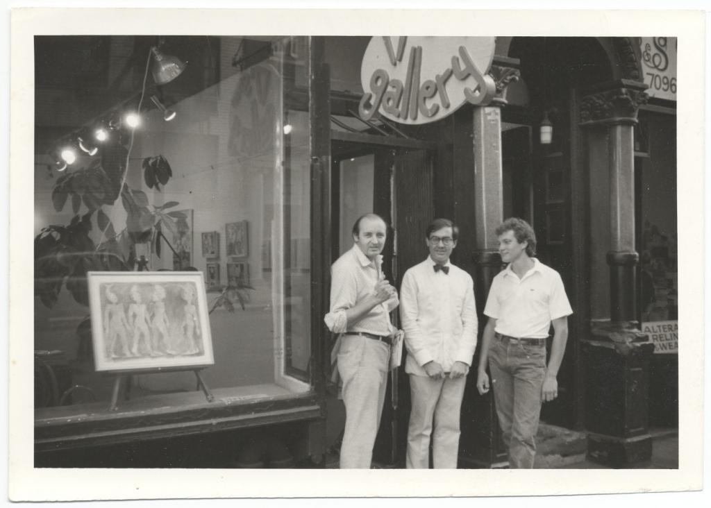 Black & white photograph of three man standing outside a gallery with columns framing the doorway and a sign above reading the name of the gallery. There is a framed painting and a plant in the window. The three man all wear white shirts of varying sleeve lengths and khakis or jeans. Giulio Blanc (left) is standing at profile view, with his head turned to the camera. N. Montero (center) is facing the camera, wearing a bowtie and glasses. The unidentified man (right) wears a white polo and faces the others. 