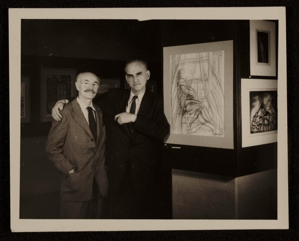 Black and white photograph of two men in a gallery. The man on the right, who is slightly taller (unidentified man), has his arms around the shoulder of the man on the left (Hugo Gellert). Both men are wearing suits and ties, and Gellert is wearing glasses and has a mustache. They are standing next to a panel with three artworks. The piece closest to them (most visible) is a charcoal line drawing of a woman sitting in a windowsill with curtains sweeping around her.