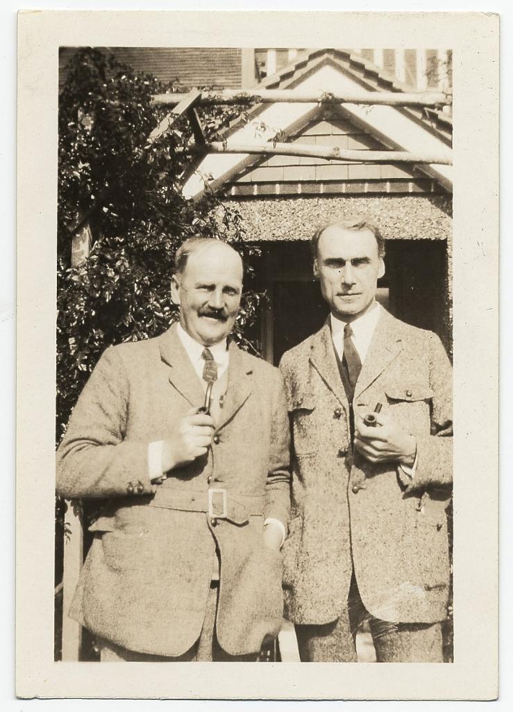 Sepia photograph of two men, both wearing formal coats and holding smoking pipes close to their chest. Kuhn, right, is slightly taller and thinner, and the unidentified man, left, has a mustache. They are standing outside in front of a house with a tree in the background.