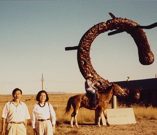 Detail of a man and woman stand in a field wearing khaki pants and light colored tops, posing for the camera. In the background a man sits on a horse wearing a cowboy hat in front of a large sculpture of a horseshoe with nails stuck in it. 