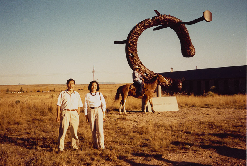 A man and woman stand in a field wearing khaki pants and light colored tops, posing for the camera. In the background a man sits on a horse wearing a cowboy hat in front of a large sculpture of a horseshoe with nails stuck in it. 