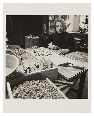 Grayscale image of Marjorie Krelick seated behind a worktable in her studio. She is wearing a black turtleneck, and there are various stones and tools spread out on the table in front of her.