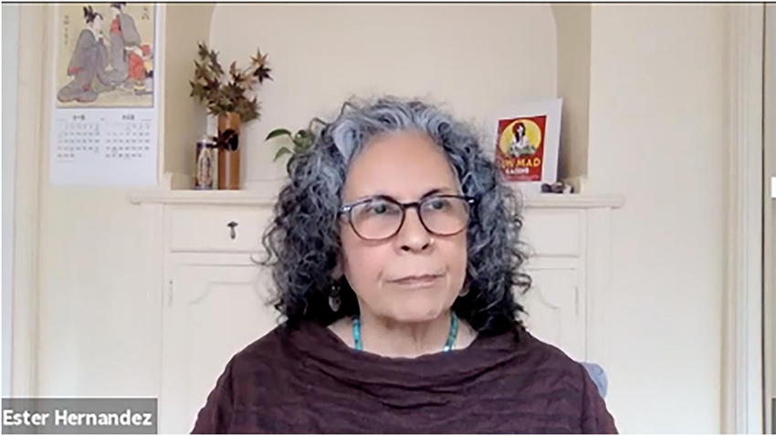 Color screenshot of Ester Hernández wearing glasses, a turquoise necklace, and a dark purple shirt, sitting in front of a white wall with a built in hutch with a calendar with a Japanese print, a vase of flowers and a print of her artwork “Sun Mad.”