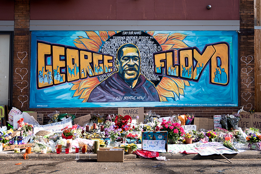 Large mural on a brick wall the words George Floyd in yellow letters against a blue background and a portrait of a man in the center against a sunflower with names listed in white lettering. There are white hearts drawn on the side of the mural and flower, candles, and protest signs laid on the ground in front of the mural.