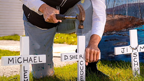 White crosses with names written in black being nailed into the grass by a kneeling woman, who is front of a church.