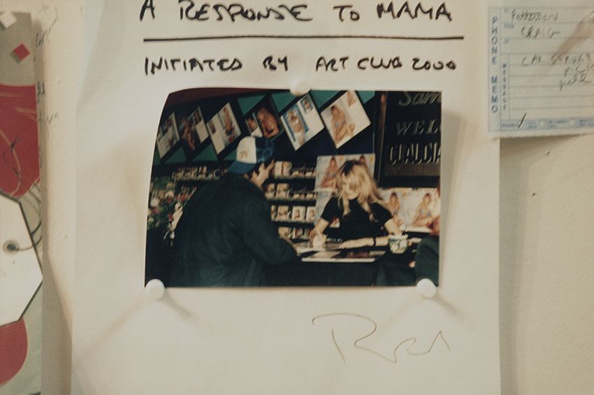 Color photograph of an artwork consisting of a piece of white paper with a photograph of Colin de Land standing at a table getting a calendar signed by Claudia Schiffer, wearing a trucker’s cap backwards. The photograph is held in place by three white plastic pushpins and has an illegible signature marking below the photograph. Above it written in capital letters in black marker is text reading “A response to MAMA initiated by Art Club 2000.” The artwork is pinned to a wall with a message from a phone memo 