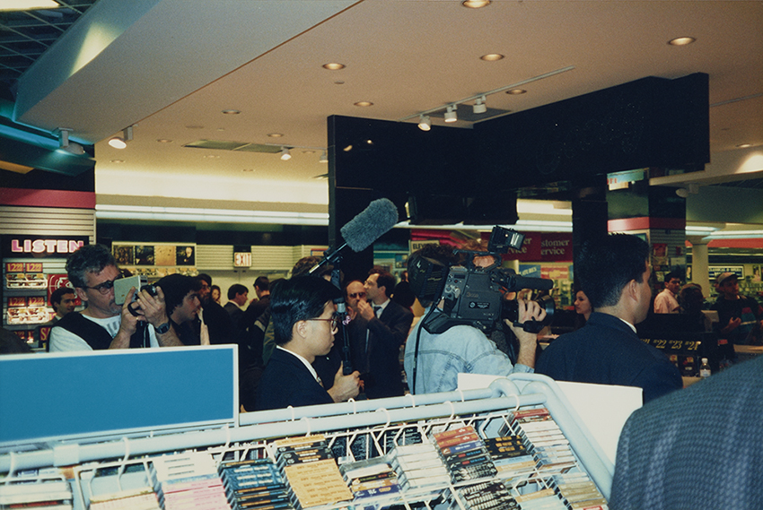Crowd at a record store event, including members of the press with recording microphones and cameras. In the foreground is a rack of cassette tapes, and a sign in the background reads LISTEN.