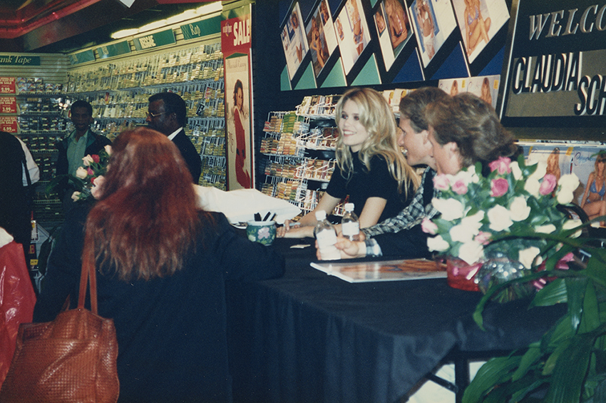 Claudia Schiffer sits at a long table covered with a black cloth with two men. There is a security guard next to the table and members of the crowd surrounding it. There are racks of cassettes and compact discs in the background, a display sign for Mariah Carey’s album Merry Christmas, and specially designed signs with pages from Schiffer’s calendar, and a sign behind the table reading Welcome Claudia Schiffer.