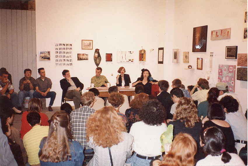 Panel discussion for group art show with several artists behind a table and sitting in cars facing an audience. Behind them are many works of art of various media hanging on white walls, and a small sculpture on a pedestal. 