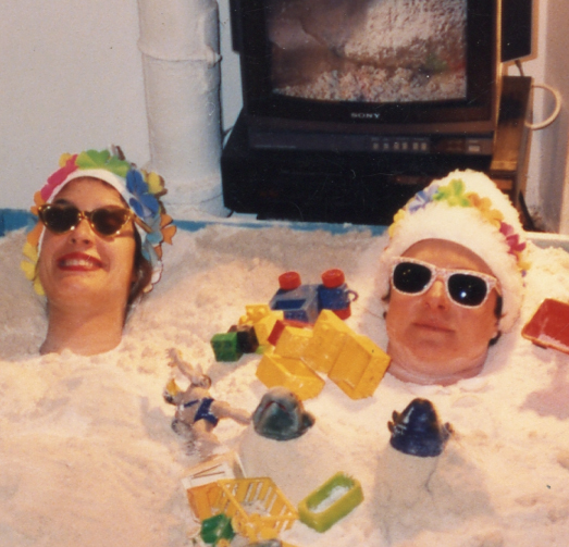 Detail of Janine Antoni and Beth Haggard buried up to their necks in a blue sandbox in a gallery setting. They are wearing sunglasses and bathing caps with rainbow-colored flowers, and there are small brightly colored toys on top of the sand. There is a light and television behind them and a person in a black t-shirt, jeans and black Chuck Taylors stands on the edge of the photograph. 