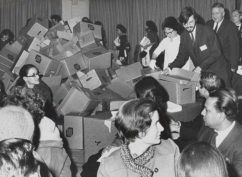 Grayscale image of many people gathered in a room , surrounding empty boxes stacked haphazardly. 