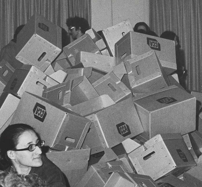 Detail of grayscale image of many people gathered in a room, surrounding empty boxes stacked haphazardly. 