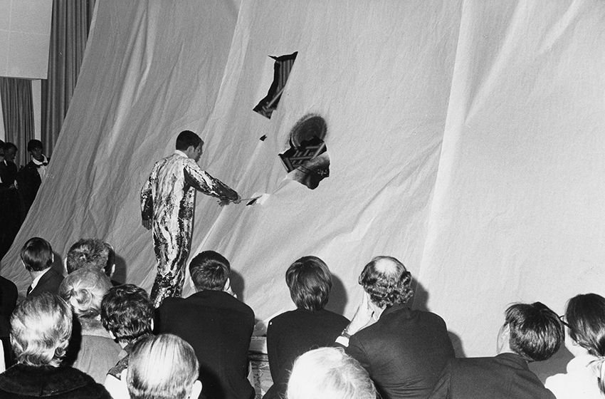 Grayscale image of a man in a paint-covered jumpsuit making holes in large scrims of paper in front of an audience.