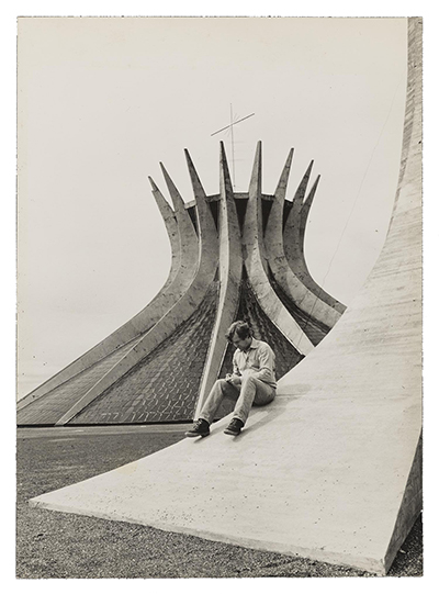 Grayscale image of Robert Hughes sitting on the sloping curve of a building examining something in his hands, with a modernist church in the background.  