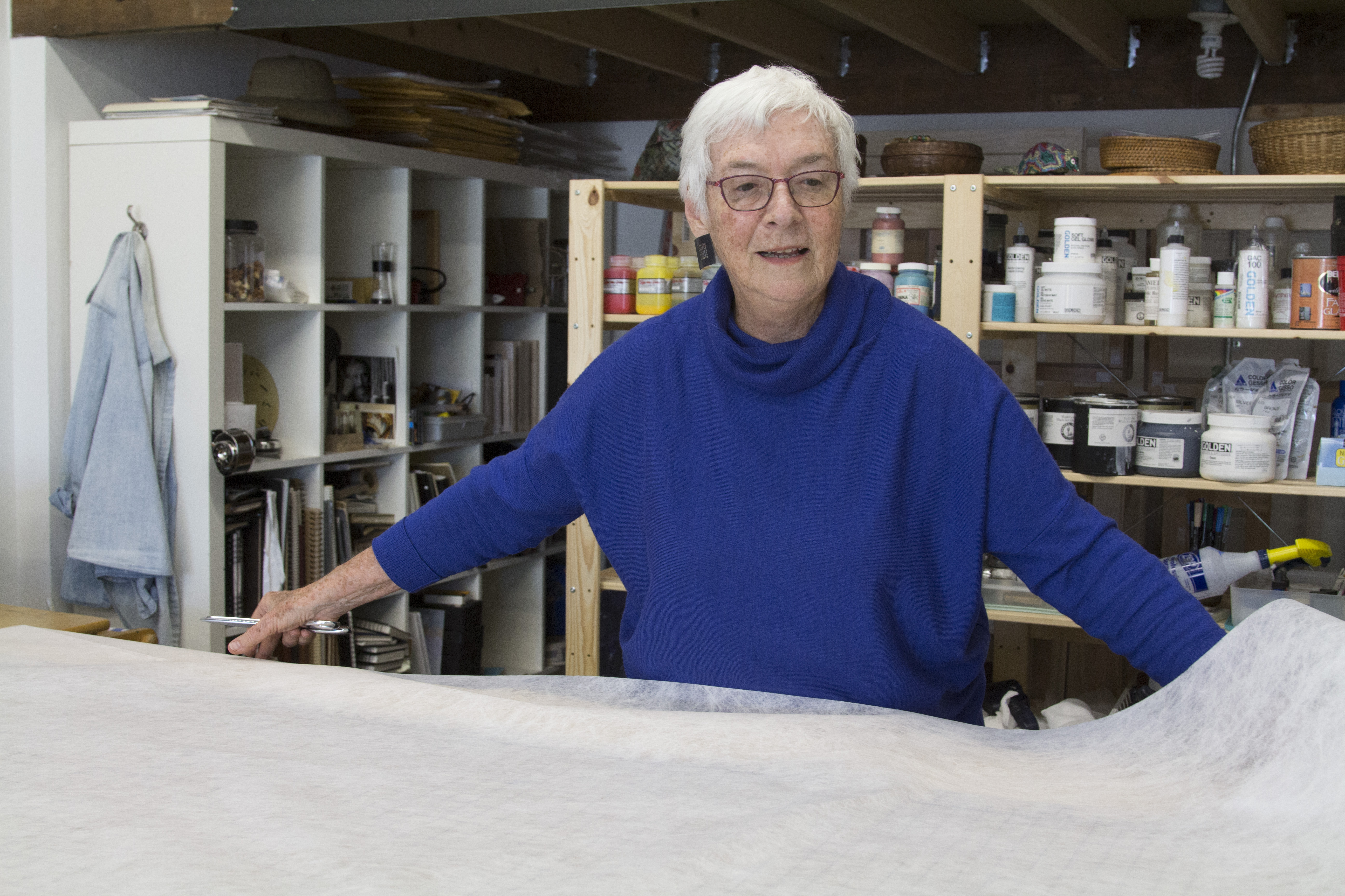 Woman with short white hair wearing a blue sweater and glasses working in an art studio.