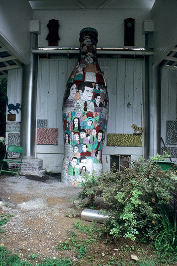 Large sculpture of a cola bottle painted with figures, standing next to a wall with other works of art displayed in a covered outdoor area with a bush in the right foreground. 