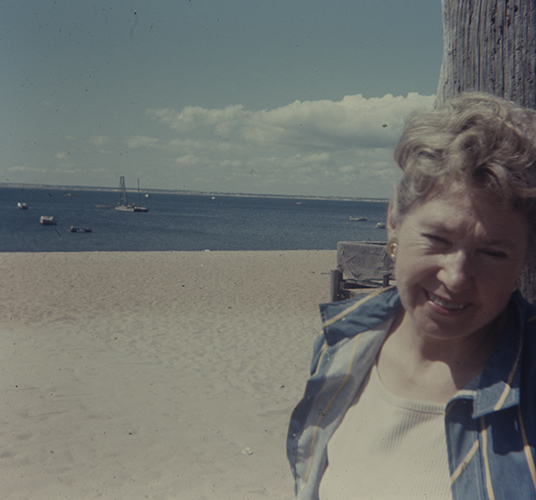 Dorothy Seckler at the beach wearing a blue plaid button down shirt and a white t-shirt. She leans against a wooden structure, with boats out in the water in the background.