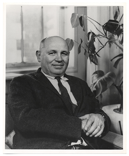 Gray scale image of Romare Bearden, in a dark suit and tie, posing for a portrait next to a plant with a window in the background.