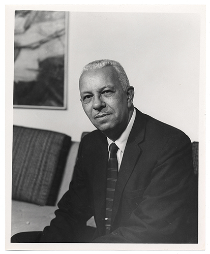 Gray scale image of Hale Woodruff posing for a portrait in a dark suit and dark striped tie. He is sitting on a light-colored sofa with a dark accent pillow with an abstract painting hanging on the wall behind him. 