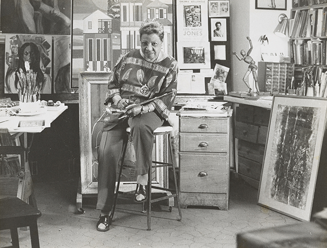 Lois Mailou Jones in her art studio surrounded by artworks, art supplies, books and ephemera. and sitting on a stool wearing pants and loose, graphic-patterned blouse.