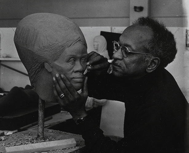 Charles White wearing a black turtleneck and wire frame glasses working on a sculpture of a head.