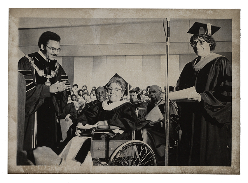 Gray scale image with Dr. James Cheek on the right applauding Alma Thomas, who sits in a wheelchair in the center, and a woman stands to her right. There is a microphone stand in the foreground of the photograph and all are wearing academic robes. 