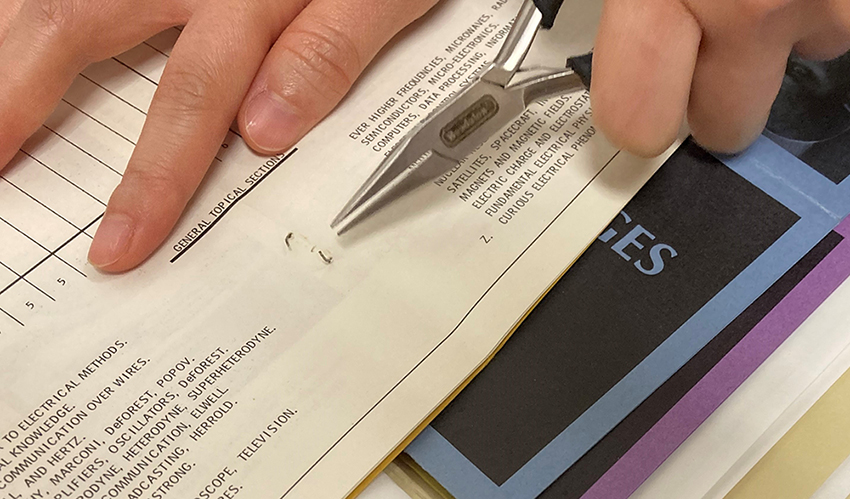 Two hands in a photograph with archival documents on white, blue, and purple paper printed in black, all in a green folder. Needle-nosed pliers are being used to remove a staple from one of the documents. 