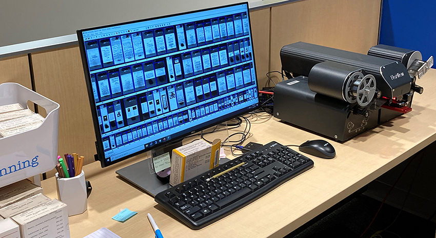 Wooden desk with black microfilm scanner, keyboard and mouse. The screen shows scans of microfilm frames, and there are boxes of microfilm, sticky notes, a blue marker on the desk, a cup with pen and markers, and white and blue walls in the photograph.