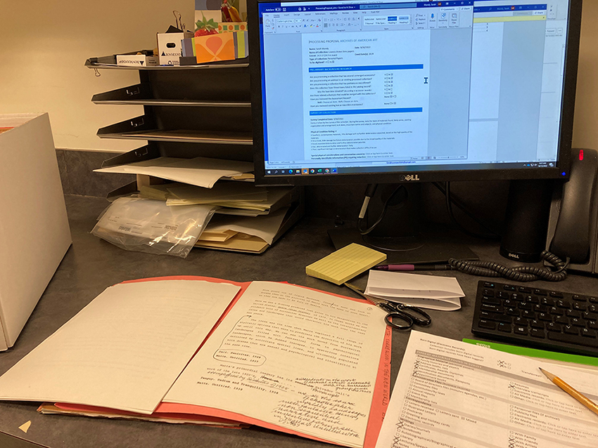 Desk with red folder open with archival materials, and another document being filled out with pencil. There is a multi-level paper tray with various supplies on it, and various supplies on the desk including scissors, sticky notes, and a telephone. The computer screen on the desk is open to electronic form. 