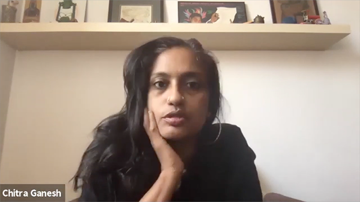 Chitra Ganesh sits below a shelf with small works of art against a white wall. She is wearing a black top and large hoop earrings and rests her chin in her right hand. 