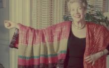 Woman draped in two different red textiles, with her arm outstretched to display one of them.