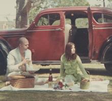 Detail of Claes Oldenburg and Patty Mucha sit on a blanket with a picnic in front of a red car with its back door open. There is a large tree in the background. 