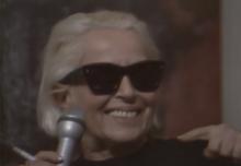 A woman with blonde hair, wearing sunglasses and a black turtleneck, holds a microphone and cigarette and smiles.