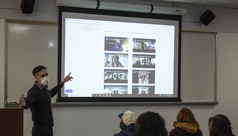 A man points to a video screen in a classroom with students seated in the foreground.